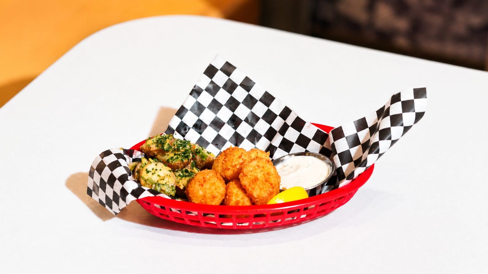 Lightly breaded scallops with tartar sauce, potato salad and a lemon wedge, in a red basket lined with black and white checked paper.