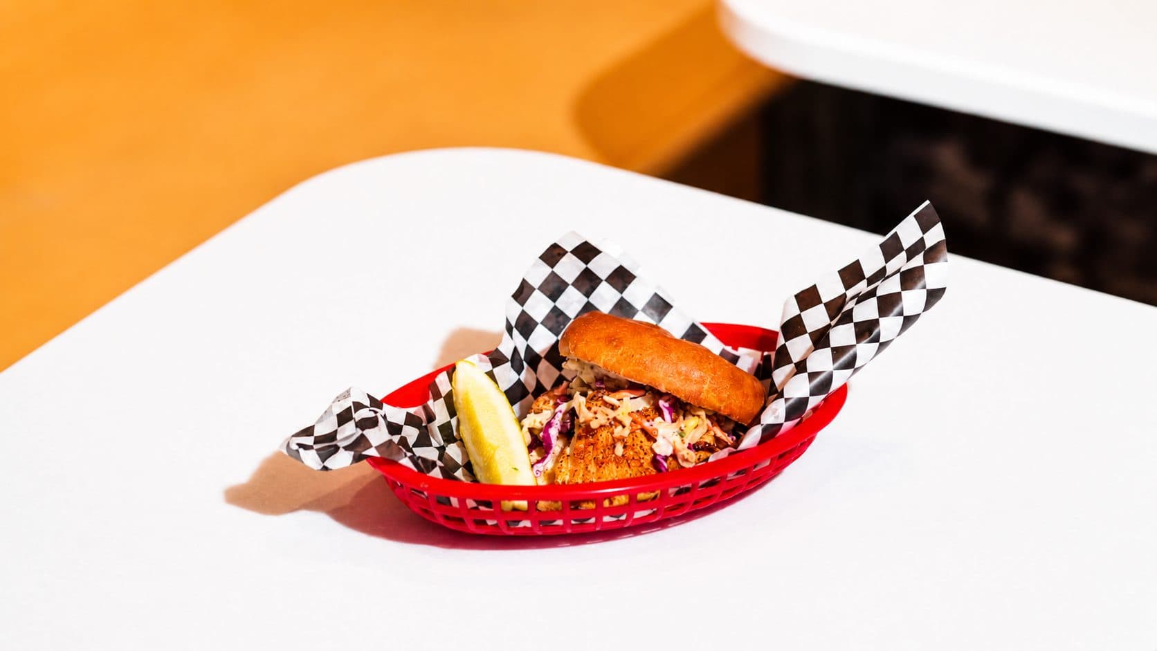 A blackened halibut cheek sandwich on a brioche bun with coleslaw and a lemon wedge, in a red basket lined with black and white checked paper.