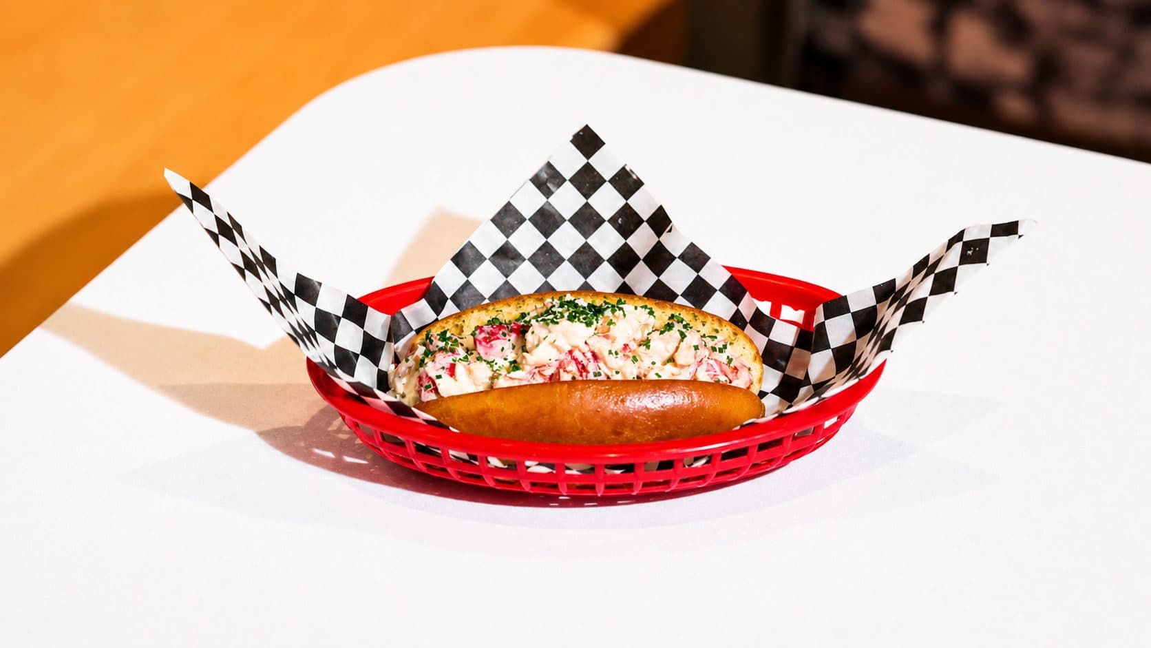 A lobster roll in a toasted split bun, piled with lobster meat and chives, in a red basket lined with black and white checked paper.