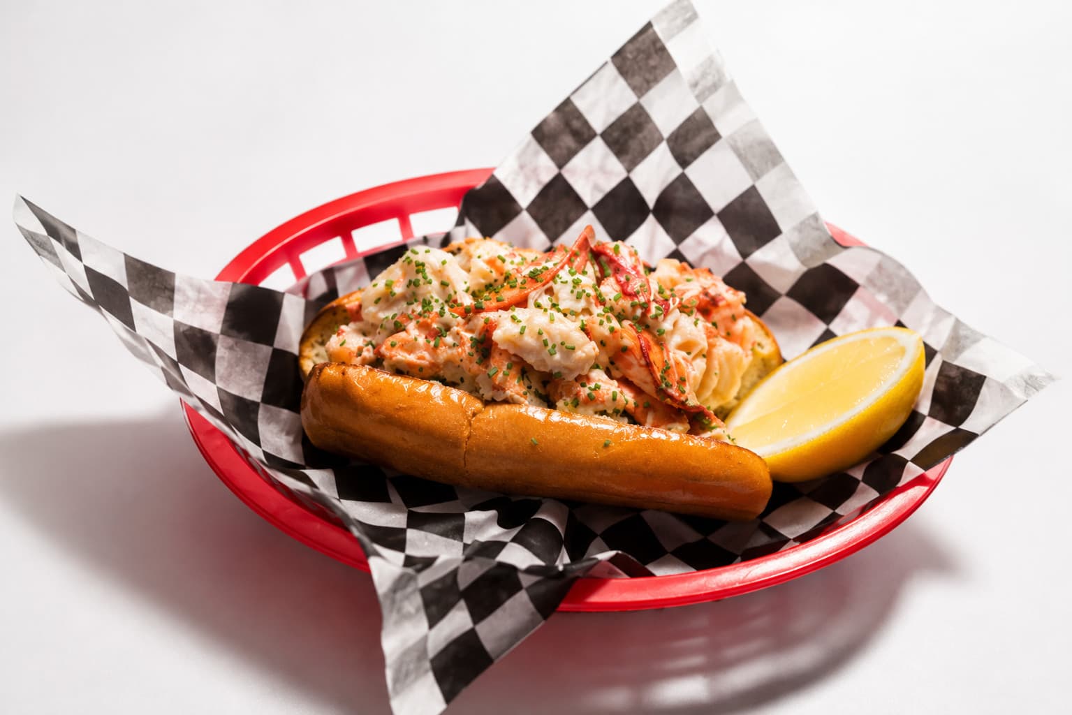 A lobster roll in a red basket lined with black and white checked paper, piled with lobster meat and chives, with a lemon wedge alongside.
